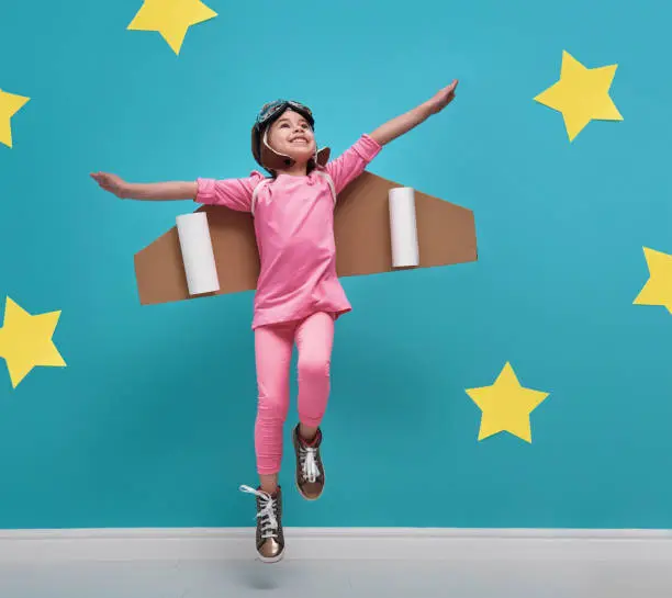 Little child girl in an astronaut costume is playing and dreaming of becoming a spaceman. Portrait of funny kid on a background of bright blue wall with yellow stars.