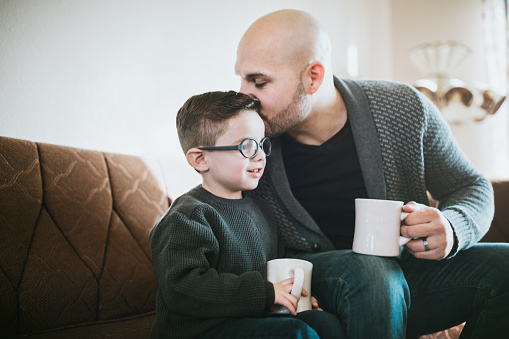 A millennial dad and his cute young boy sit together in a sun lit morning living room enjoying a mug of coffee or hot chocolate.  A depiction of a loving, caring, and supportive father.