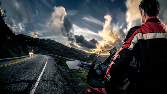 Male motorcyclist wearing protective leather racing suit with a red bike or motorcycle on an open road.  The image is shot in HDR and composite.  The vehicle is cropped to become generic non branded. The image depicts travel and adventure.
