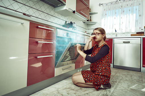 young woman inside her kitchen opening the stove, burning food inside it, negative expression.