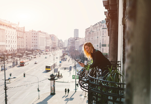 Single woman drinking coffee and using her smart phone on hotel balcony