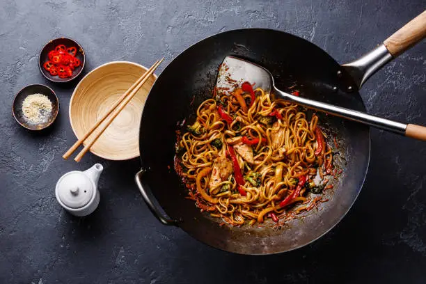Udon stir-fry noodles with chicken and vegetables in wok pan on dark stone background