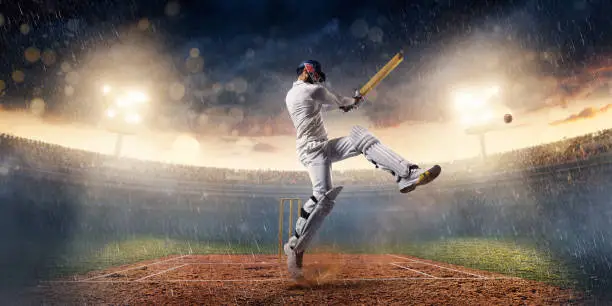 Cricket batsman bouncing a ball. He is wearing unbranded sports cloth and equipment. The bleachers full of people are blurred behind the player. There is intentional lenseflares on the image. The stadium is made in 3D.