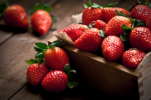 Small wooden crate filled with fresh organic strawberries on rustic wood table. Three fruits are out of the crate. Predominant colors are red and brown. Selective focus. Low key DSRL studio photo taken with Canon EOS 5D Mk II and Canon EF 70-200mm f/2.8L IS II USM Telephoto Zoom Lens