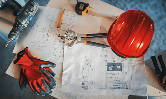 Closeup top view of typical construction worker tools and protective gear. There are blueprints, electric drill, tape measure, clamps, gloves and a hardhat.