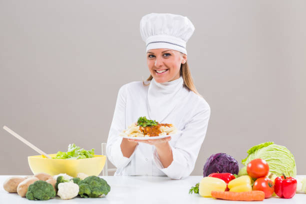Cheerful female chef is sitting at the table with bunch of vegetable and showing prepared meal.