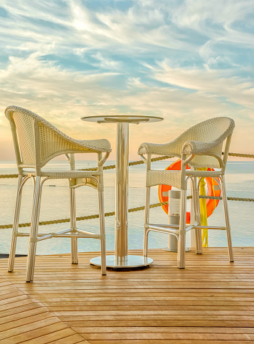 Bar table and two bar stools made of rattan on the wooden deck amid the picturesque sunset