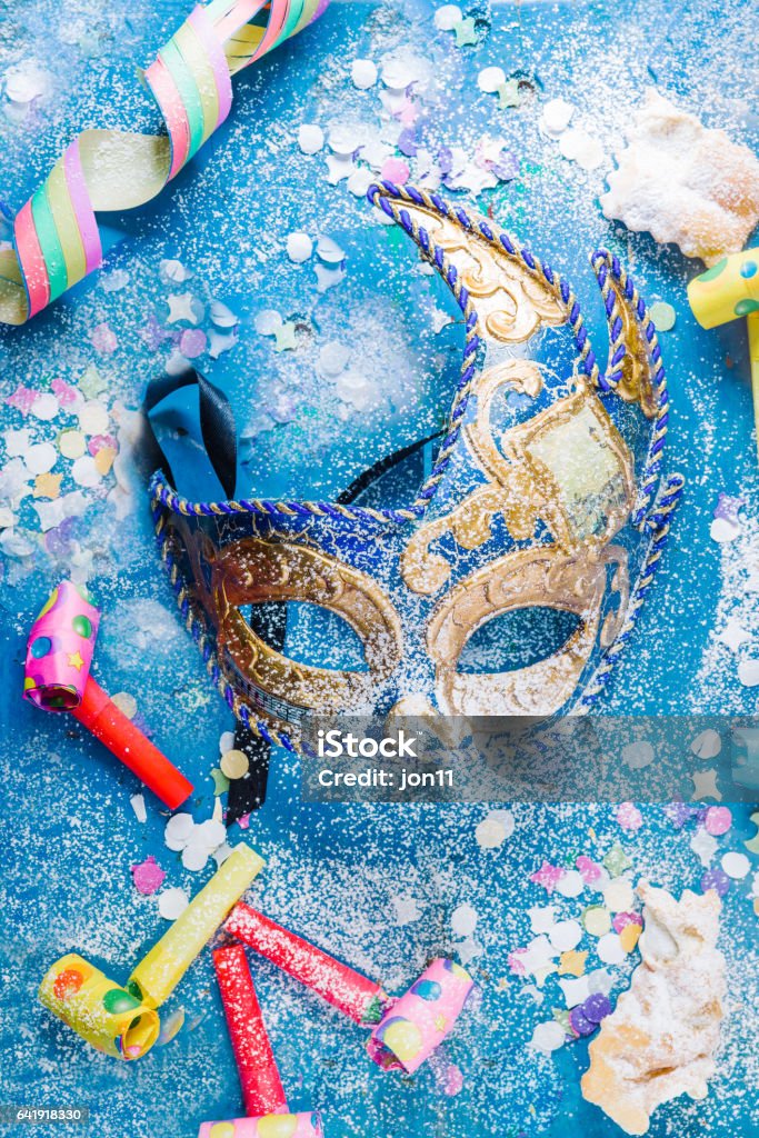 Carnival time! Carnivalesque composition. Top view of a carnival mask, whistles, corianders, and sfrappoles or chiacchere,italian fried carnival cookies with icing sugar on a bluish shabby chic board. Plate Stock Photo