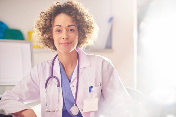 female doctor portrait a female doctor sits at her desk and smiles proudly to camera.She is wearing blue scrubs and a lab coat . in pride we trust stock pictures, royalty-free photos & images