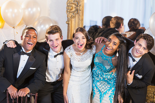 A group of five multi-ethnic teenagers and  young adults dressed in formalwear - dresses and tuxedos. They are at a special event, a party or prom, standing together in a row, laughing and smiling.