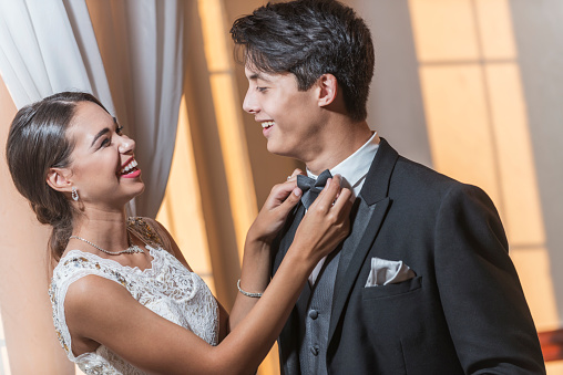A teenage couple wearing formal attired, going to the prom or other special formal event. The mixed race young woman is adjusting her date's bowtie.