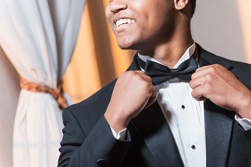 Cropped view of a young black Hispanic man wearing a tuxedo. He is smiling, dressed for a special event, perhaps a wedding or prom. He is adjusting his bowtie.