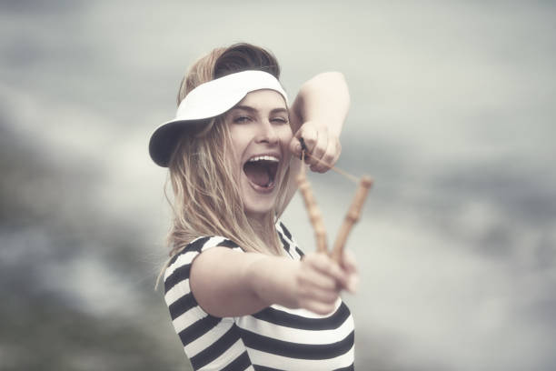 funny young woman aiming with the slingshot to the camera, laughing and enjoying playing with it.