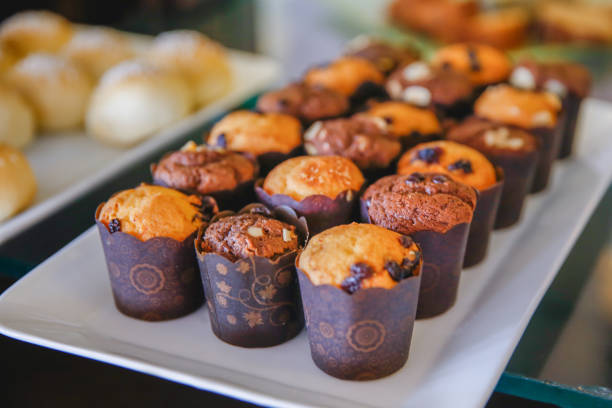 Chocolate muffin with raisin and almond slice on top. The muffin has a brown cover paper. There's collection of bread on the blurred backgrounds.