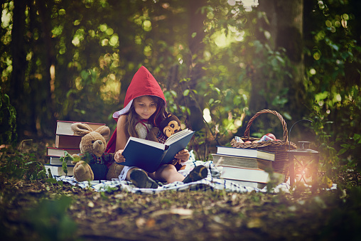 Shot of a little girl in a red cape reading a book with her toys in the woods