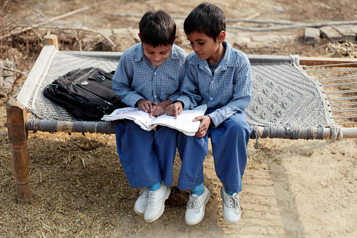-      Two rural children sitting on a cot and reading a book carefully.