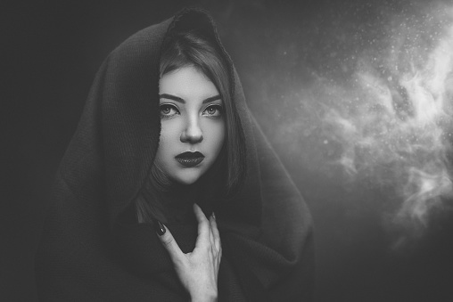 monochrome shot of woman wearing hood, being terrified hiding and looking at camera, smoke background behind her.