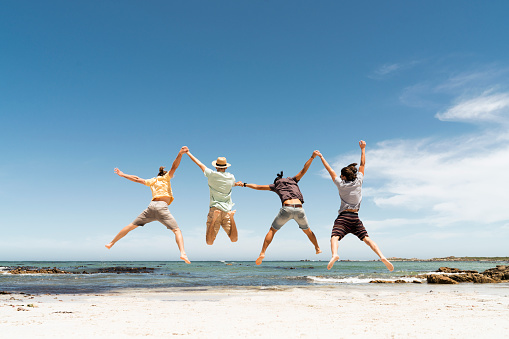 A photo of friends jumping on shore. Full length rear view of men are holding hands. They are in casuals while enjoying summer vacation at beach.