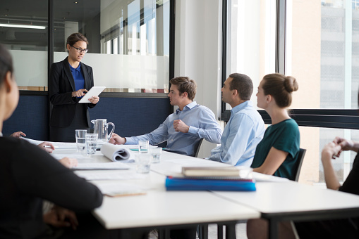 Confident businesswoman using digital tablet while giving presentation to colleagues in office. Female professional is discussing strategy with executives in board room. All are in meeting at workplace.