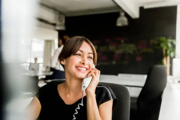 Cheerful businesswoman using telephone in office. Happy female professional is looking away while sitting on chair. Beautiful executive is at brightly lit workplace.