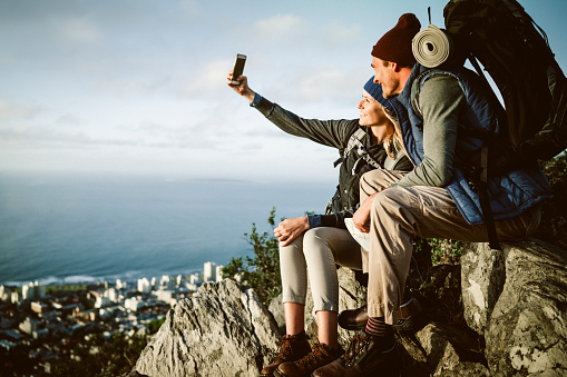 Girlfriend taking selfie with boyfriend on mountain. Backpackers enjoying vacation. They are sitting on rocks overlooking cityscape and sea.