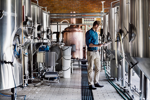 Male worker using digital tablet in brewery. Full length of professional in formals is working amidst metallic vats. He is in beer industry.