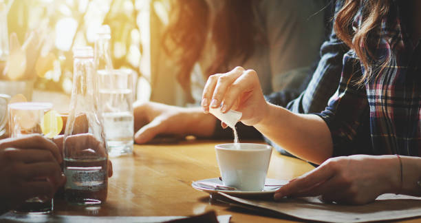 Closeup of unrecognizable woman putting some sugar into her coffee. She's having a tea spoon of sugar packed in a little bag. Blurry people in background.