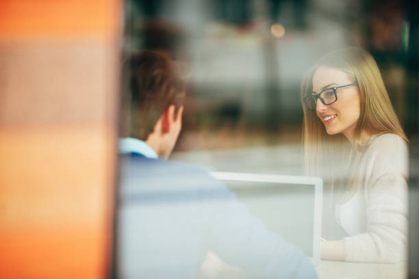 Beautiful young woman smiles and talks to a young guy at cafe. Both are casually dressed and there is a laptop beside them. It seems that they are having fun. Shoot through the window, over the male shoulder. selective focus, copy space has been left.