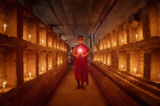 Young burmese buddhist novice monk standing in the middle of a tomb inside temple pagoda surround with burning candles, praying and worshipping. Real People Portrait. Old Bagan, Myanmar, South East Asia.