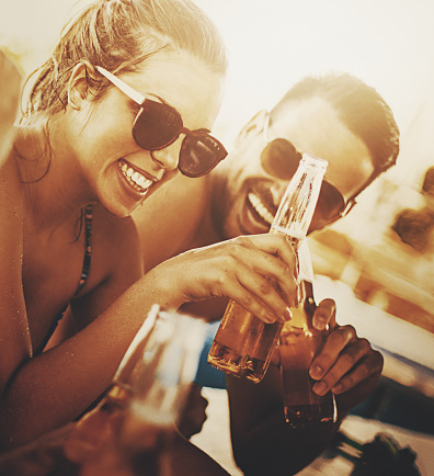 Closeup front view of couple of young adults having fun by a poolside bar on a hot summer afternoon. They are drinking beers and laughing, sunshine flaring from the background.