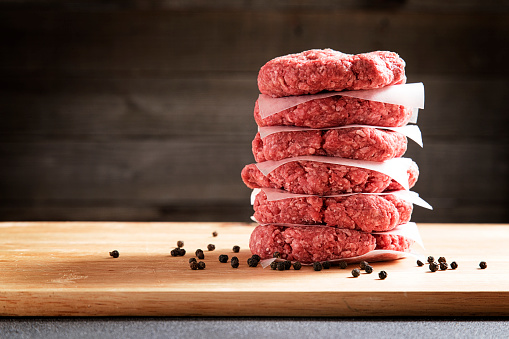 stack of six raw hamburgers on wax paper on wood cutting board ready to be grilled with uncracked peppercorns and copy space to left
