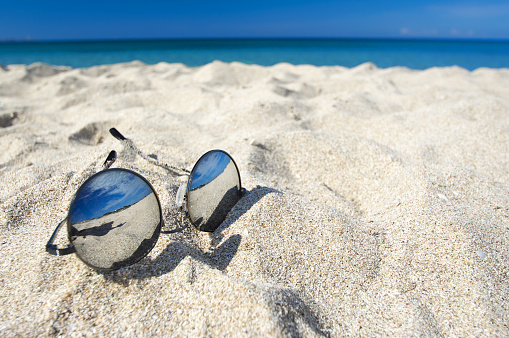 Round sunglasses with mirror reflection in white sand on sunny summer beach.