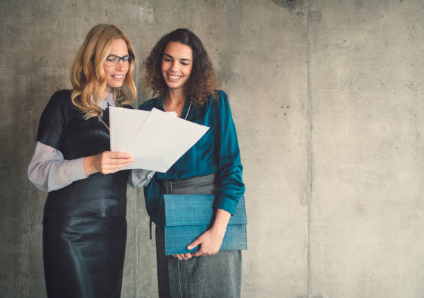 Two girls in the office with folders with papers in hand. Young women discuss office work. Blue empty hall on the background