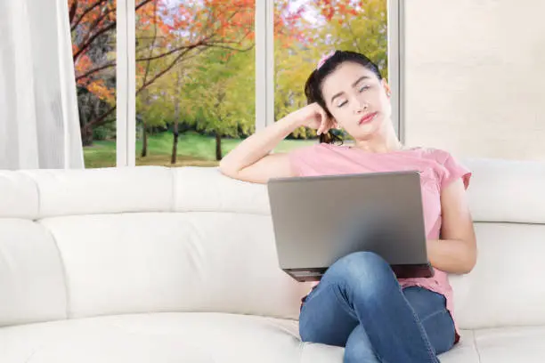 Portrait of a female model sitting on the couch and looks sleepy with a laptop computer, shot with autumn background on the window
