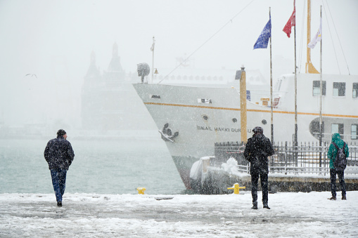 People are watching the scenic at Kadikoy in a snowy day with a traditional public transportation ship and The Haydarpasa Train Station Building at the background.