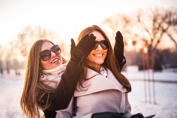 One friend suprising another from the back by covering her eyes in the park on sunny winter day