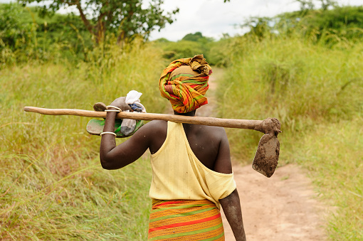 African woman with the turban on the head going dirt road to work with the hoe in field in Tanzania