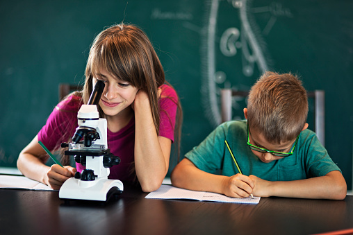 Primary school biology lesson. Girl and boy sitting at the desk and using microscope. Both are making notes while the girl is looking into the microscope eyepiece
