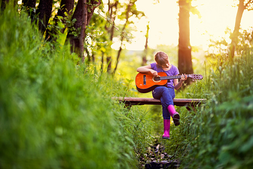 Little girl aged 9 is sitting on the small bridge in the forest. The girl is playing a guitar. Sunny spring evening.\n