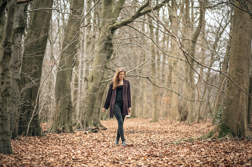 A beautiful young woman in her early 20s walking through a forest with a leaf-strewn floor.