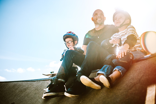 A fun, playful Hispanic Dad and his two boys play together at a skate park. They boys have helmets and padding, and are ready to have fun with their father. The young family of three takes a minute to rest together. A sun flare shines behind them. Horizontal with copy space.