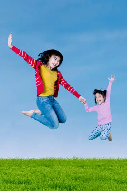 Mother and cute daughter jumping together on the air while raising hands under the clear sky