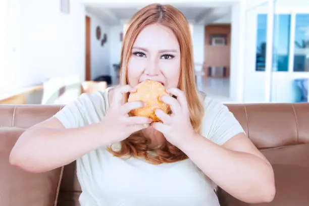 Image of a beautiful blonde woman with overweight body sitting on the sofa while eating hamburger