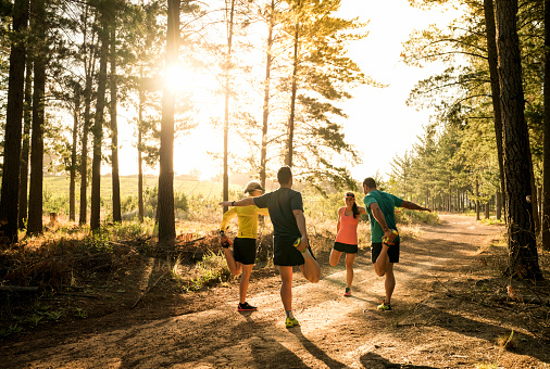 A photo of male and female friends stretching legs before jogging. Full length of people wearing sportswear on dirt road. They are exercising in nature.