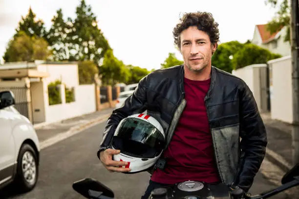 Portrait of confident man sitting on motorcycle. Mature male is holding crash helmet. He is wearing leather jacket on street.