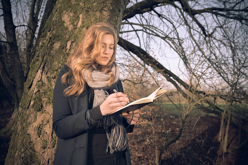A beautiful young woman in her early 20s and dressed in black and grey stands by a tree in woodland whilst writing in her journal on a winter day.