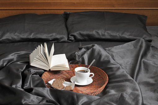 Cup of coffee with milk chocolate on wicker tray and open book in bed with black satin linen and wooden bedhead. Text in book unreadable.