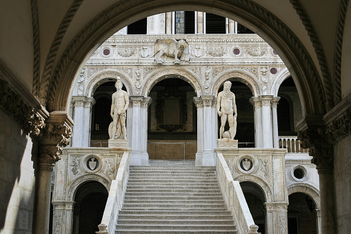 Doge's Palace  and Giants’ Staircase as seen from Courtyard, Venice, Italy. The Scala dei Giganti (Giants’ Staircase) is flanked by Mars and Neptune and framed by an arch. The Doge's Palace (Italian: Palazzo Ducale) was built in Venetian Gothic style in Piazza San Marco. The palace was the residence of the Doge of Venice, the supreme authority of the former Republic of Venice. It opened as a museum in 1923. Vivid blue sky is in background.