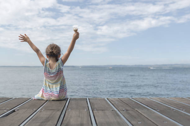 A little girl sit on a pier and is happy about her ice. She is rising up the Arms