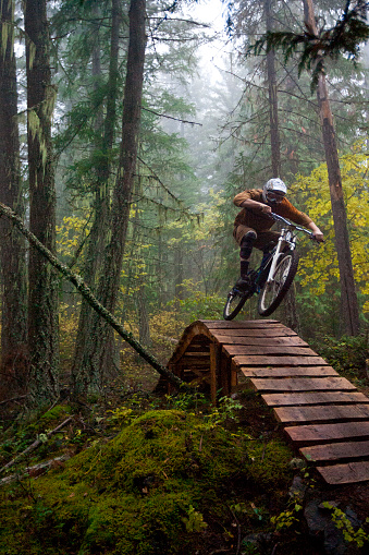 A teenager rides his downhill mountain bike off a wooden ladder bridge in a foggy forest in the fall.